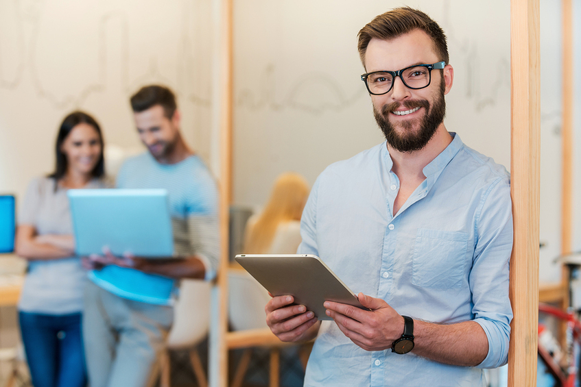 Man holding a table smiling with a couple in the background looking at a laptop.